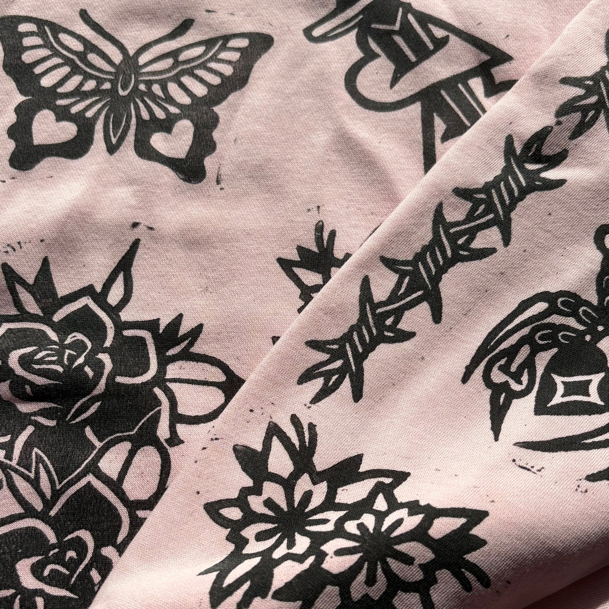 A close-up of a light pink hoodie featuring a black tattoo-style graphic print with butterflies, roses, and a dagger motif.