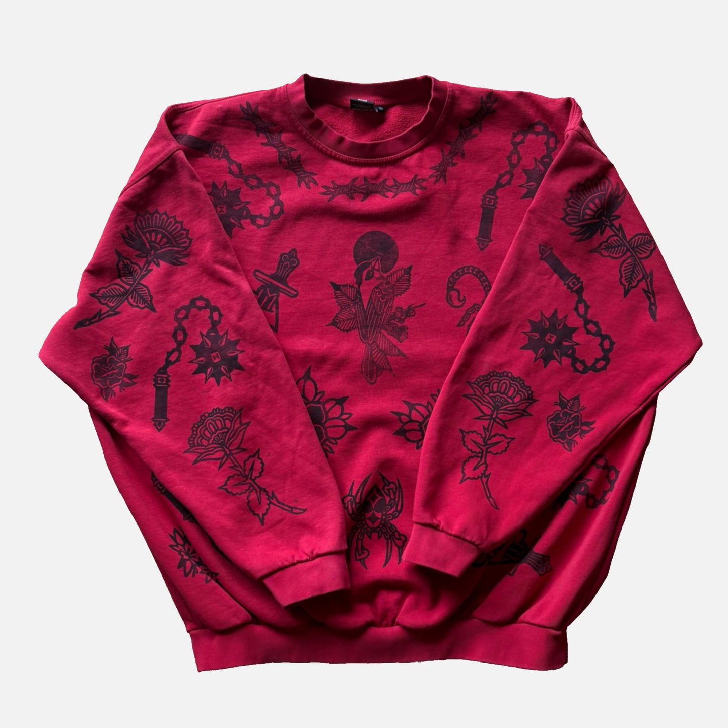 A red crewneck sweatshirt featuring an all-over black graphic print of roses, chains, and gothic symbols, laid flat against a light gray background.