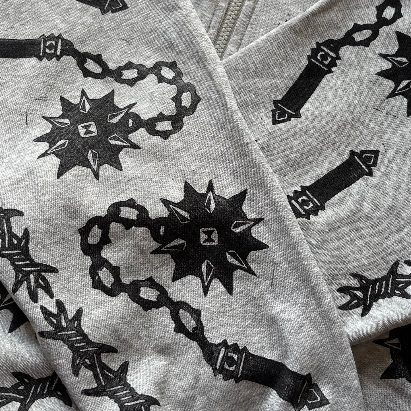 A close-up of a light gray zip-up hoodie featuring a black graphic print of flails and barbed wire, laid flat.