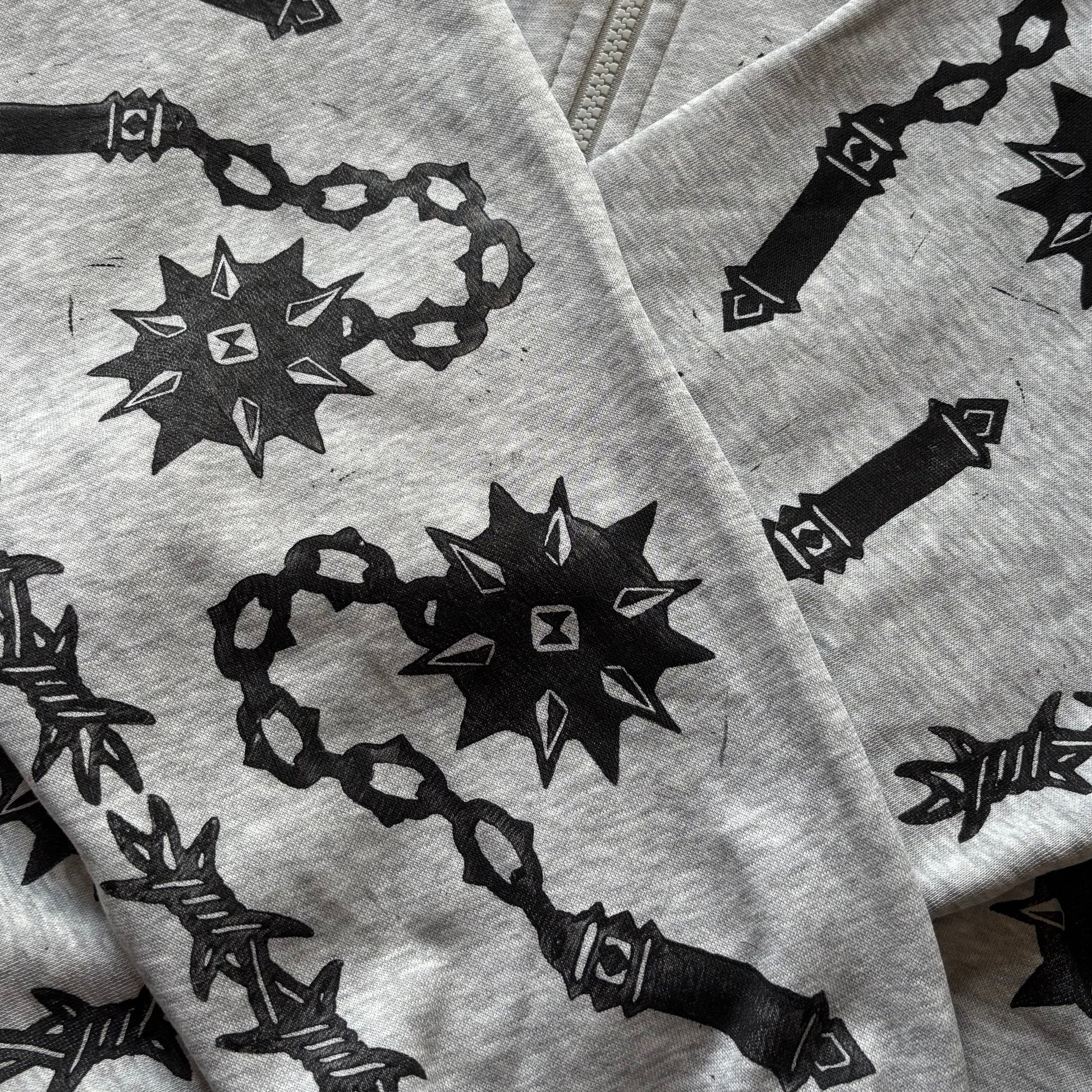 A close-up of a light gray zip-up hoodie featuring a black graphic print of flails and barbed wire, laid flat.