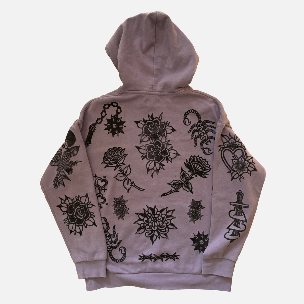 A light purple/lavender hoodie shown from the back, featuring black tattoo-style illustrations of roses, scorpions, hearts, and other floral motifs across the back and sleeves.