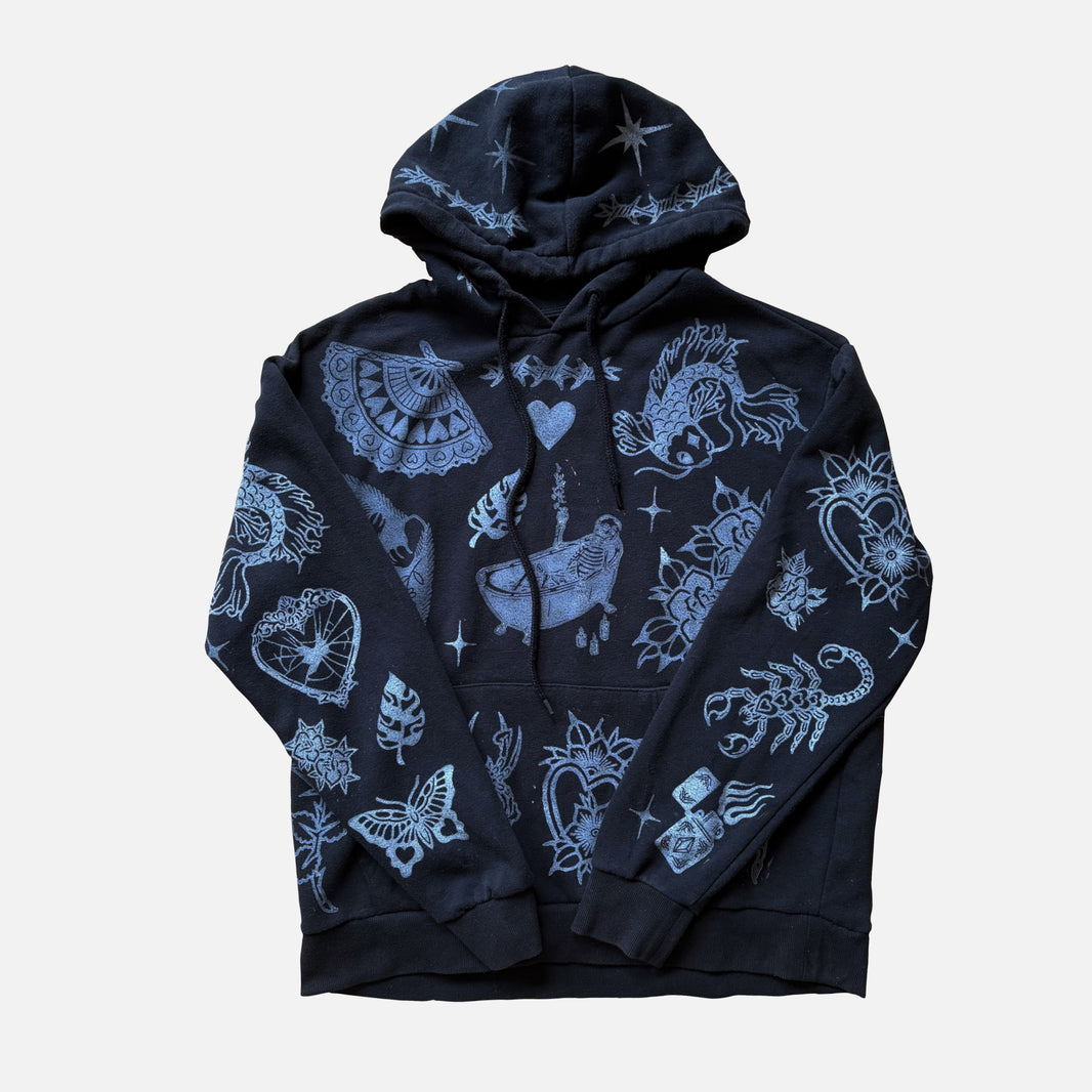A black pullover hoodie with a white graphic print featuring various tattoo-style designs including a scorpion, butterfly, heart, and celestial motifs, displayed against a light gray background.