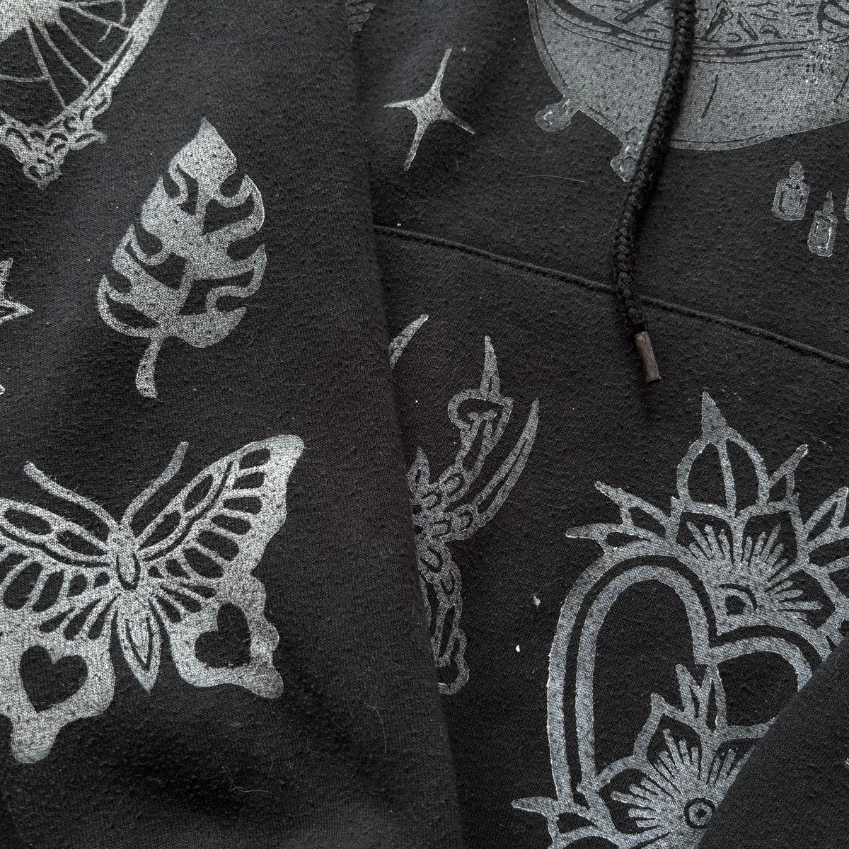 Close-up of a black hoodie featuring a white graphic print of a butterfly, leaf, spider, and floral motifs.