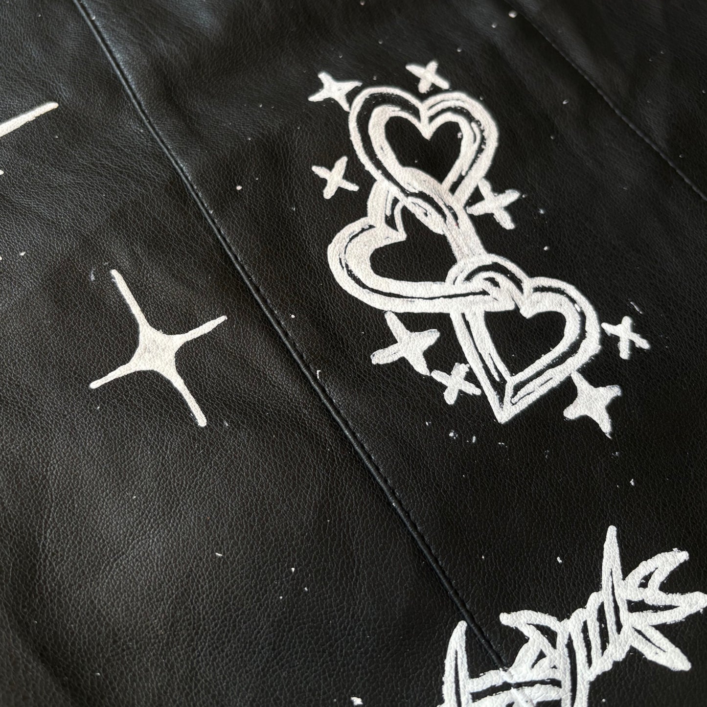 Close-up of a black textured leather mini skirt featuring hand-painted white graphics, including a triple heart design with stars and barbed wire.